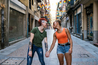 Coole junge homosexuelle Frauen mit Sonnenbrillen und Tattoos, die sich gegenseitig ansehen, während sie die Zunge herausstrecken und sich auf einem Gehweg in der Stadt an den Händen halten - ADSF31029