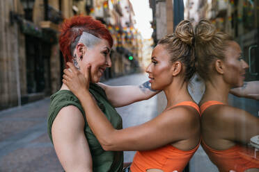 Seitenansicht eines trendigen, fröhlichen, jungen, lesbischen Paares mit Tattoo, das sich umarmt und sich im Moment des Kusses an eine Mauer in der Stadt lehnt - ADSF31031