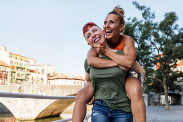 Positive lesbische Frau gibt fröhliche geliebte Huckepack Fahrt, während Spaß gegen Brücke und städtischen Gebäuden haben - ADSF31034