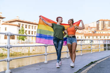 Cool tattooed woman with mohawk and LGBTQ flag embracing girlfriend with closed eyes against canal in town - ADSF31036