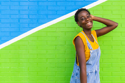 Seitenansicht einer glücklichen jungen Afroamerikanerin, die lächelnd auf einer bunten hellen Wand steht - ADSF31039