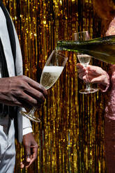 Crop anonymous woman pouring champagne from bottle into glass of black boyfriend during New Years Eve celebration against tinsel - ADSF31049