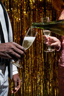 Crop anonyme Frau gießt Champagner aus der Flasche in das Glas des schwarzen Freundes während der Silvesterfeier gegen Lametta - ADSF31049