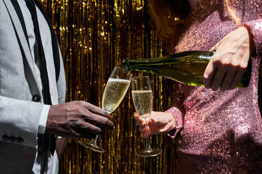 Crop anonyme Frau gießt Champagner aus der Flasche in das Glas des schwarzen Freundes während der Silvesterfeier gegen Lametta - ADSF31050