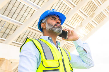 From below middle aged Hispanic foreman in in hardhat and waistcoat speaking on smartphone looking away while standing near solar power station - ADSF31064