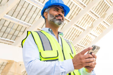 From below middle aged Hispanic foreman in in hardhat and waistcoat browsing on smartphone looking away while standing near solar power station - ADSF31065