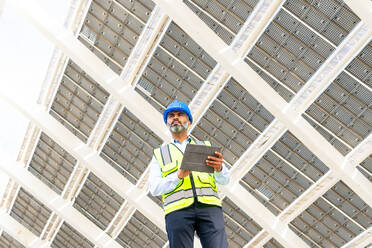 Low angle of middle aged Hispanic male engineer in uniform with tablet standing looking away under solar power station - ADSF31066