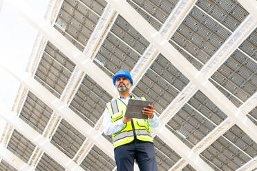 Niedriger Winkel eines männlichen Ingenieurs mittleren Alters in Uniform mit Tablet, der unter einem Solarkraftwerk steht und wegschaut - ADSF31066
