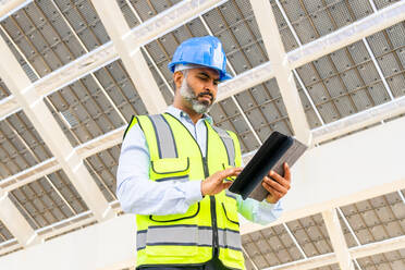 Low angle of middle aged Hispanic male engineer in uniform with tablet standing looking at screen under solar power station - ADSF31067