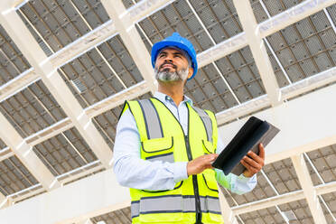 Niedriger Winkel eines männlichen Ingenieurs mittleren Alters in Uniform mit Tablet, der unter einem Solarkraftwerk steht und wegschaut - ADSF31068