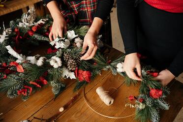 Unkenntlich gemachte Freundinnen stehen an einem Holztisch und basteln einen kreativen Weihnachtskranz aus dekorativen Tannen und roten Bändern für die Weihnachtsfeier - ADSF31101