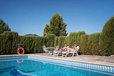 Carefree senior couple in swimwear and sunglasses sunbathing on sunbeds with drinking coffee near pool and hedge - ADSF31126