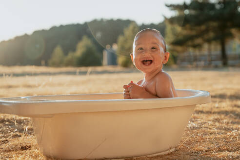 Seitenansicht eines glücklichen Kleinkindes mit Spielzeug, das in einer Plastikwanne sitzt und wegschaut, während es mit Wasser in der Landschaft spielt - ADSF31129