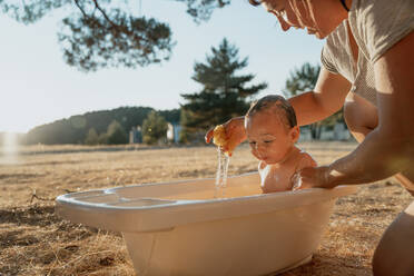 Cropped unrecognizable mom bathing toddler child while sitting on meadow under shiny sky on sunny day - ADSF31130