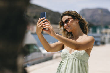 Lächelnde Frau mit Sonnenbrille nimmt Selfie durch Smartphone - ABZF03597