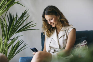 Smiling woman text messaging through mobile phone while sitting on sofa - ABZF03607