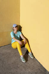 Frau mit Mütze sitzt an einer gelben Wand an einem sonnigen Tag - VPIF05065