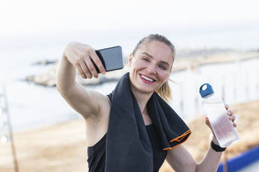 Sportswoman taking selfie through smart phone while holding water bottle - PNAF02376