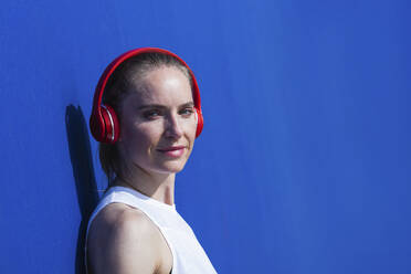 Mid adult female athlete with headphones near blue wall - PNAF02391