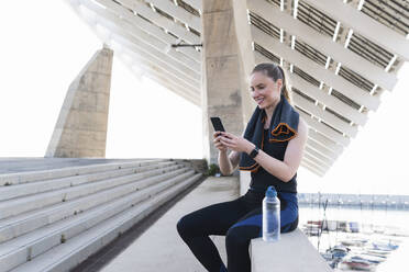 Lächelnde Sportlerin, die ihr Smartphone benutzt, während sie auf einer Stützmauer sitzt - PNAF02400