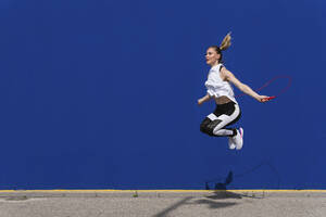 Weibliche Athletin beim Seilspringen vor einer blauen Wand - PNAF02423