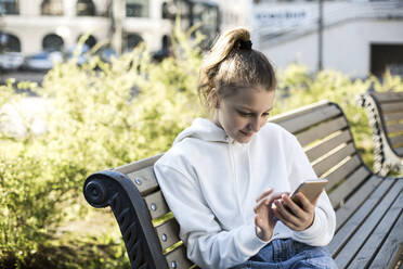 Mädchen, das ein Smartphone benutzt, während es auf einer Bank sitzt - LLUF00134