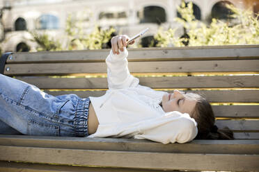 Mädchen nimmt Selfie durch Smartphone, während auf der Bank liegend - LLUF00136