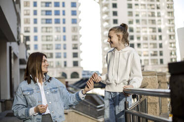Mother giving mobile phone to daughter standing at railing - LLUF00141