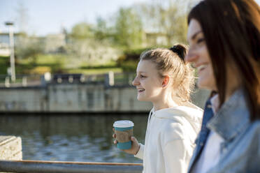 Lächelndes Mädchen hält Kaffeetasse beim Spaziergang mit Mutter am Ufer - LLUF00145