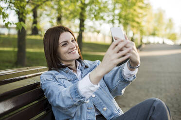 Lächelnde Frau nimmt Selfie durch Handy auf Bank - LLUF00149