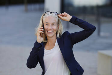 Lächelnde Geschäftsfrau, die mit einem Smartphone telefoniert, während sie die Brille auf dem Kopf verstellt - AANF00118