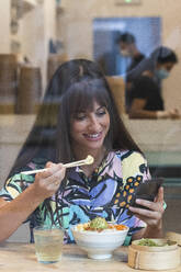 Woman using mobile phone while having food at restaurant - PNAF02470