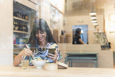 Smiling woman with smart phone having food at restaurant seen through glass - PNAF02471