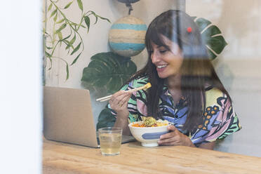 Frau, die an einem Videoanruf über einen Laptop teilnimmt, während sie am Restauranttisch isst - PNAF02472