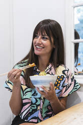Smiling woman holding bowl while eating poke at restaurant - PNAF02480