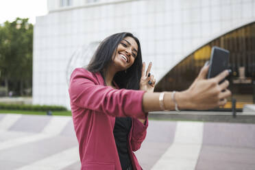 Glückliche Geschäftsfrau gestikuliert Friedenszeichen bei der Aufnahme von Selfie durch Smart-Phone - JCCMF04170