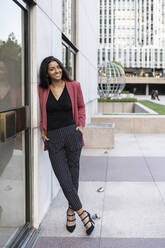 Young businesswoman standing with hands in pockets leaning on building - JCCMF04197