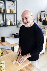 Happy senior man cutting banana in kitchen - GIOF13782