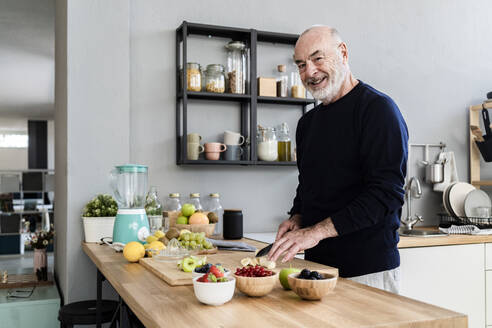 Smiling senior man cutting fruits in kitchen at home - GIOF13783