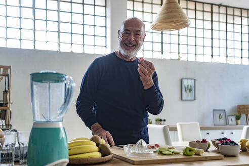Happy senior man holding slice of fruit in kitchen - GIOF13785