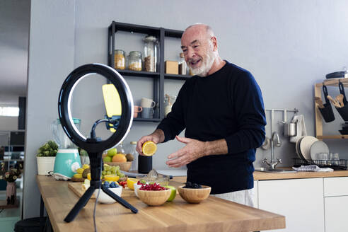 Männlicher Influencer mit Zitrone, der an der Küchentheke mit dem Handy filmt - GIOF13791