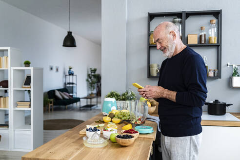 Smiling man using smart phone while preparing smoothie at home - GIOF13800