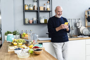 Happy man using mobile phone while leaning on kitchen counter - GIOF13801