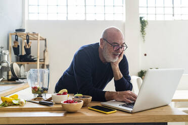 Senior man using laptop while leaning on kitchen counter - GIOF13811