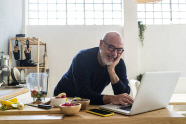 Smiling man with laptop leaning on kitchen counter - GIOF13812