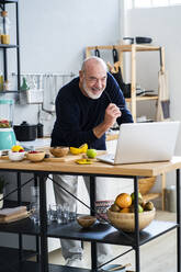 Cheerful senior man using laptop at kitchen counter - GIOF13818