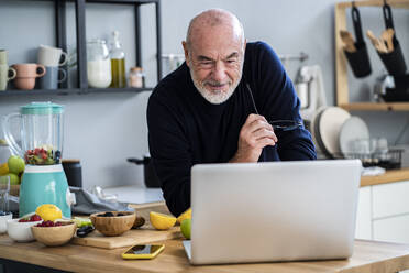 Älterer Mann schaut auf einen Laptop, während er sich auf den Küchentisch stützt - GIOF13819