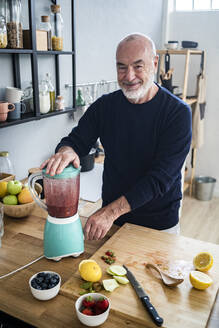 Smiling man preparing smoothie in electric juicer at kitchen counter - GIOF13821