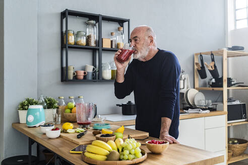 Man drinking smoothie while standing at kitchen counter - GIOF13828