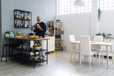 Älterer Mann hält ein Glas Smoothie in der Hand, während er sein Smartphone zu Hause benutzt - GIOF13832